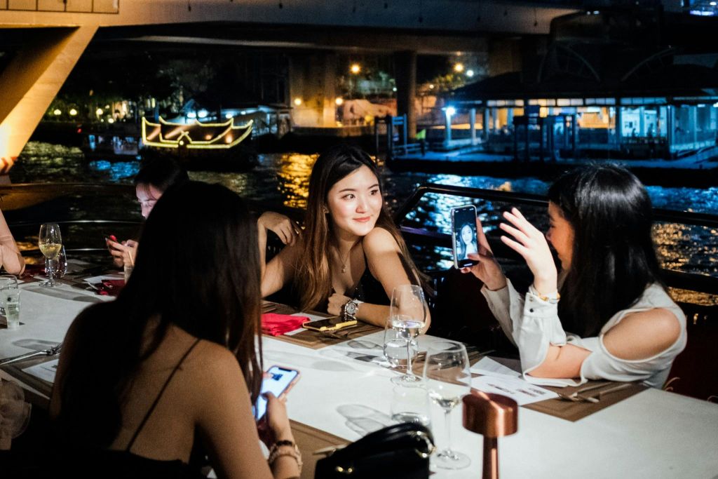 밤에 야외 레스토랑 테이블에 앉아 있는 네 명의 여성. 한 여성이 휴대폰 화면을 다른 여성에게 보여주고 있습니다. 물 위를 배경으로 보트에 불을 붙이세요.