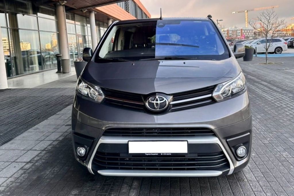 건물과 보도를 배경으로 야외 도심에 주차된 회색 도요타 밴의 전면 모습.