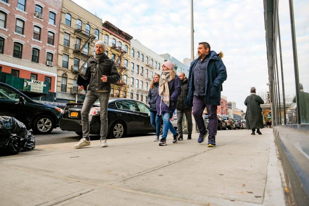 겨울 옷을 입고 주차된 자동차와 건물을 배경으로 도시 보도를 걷는 네 명의 사람들.