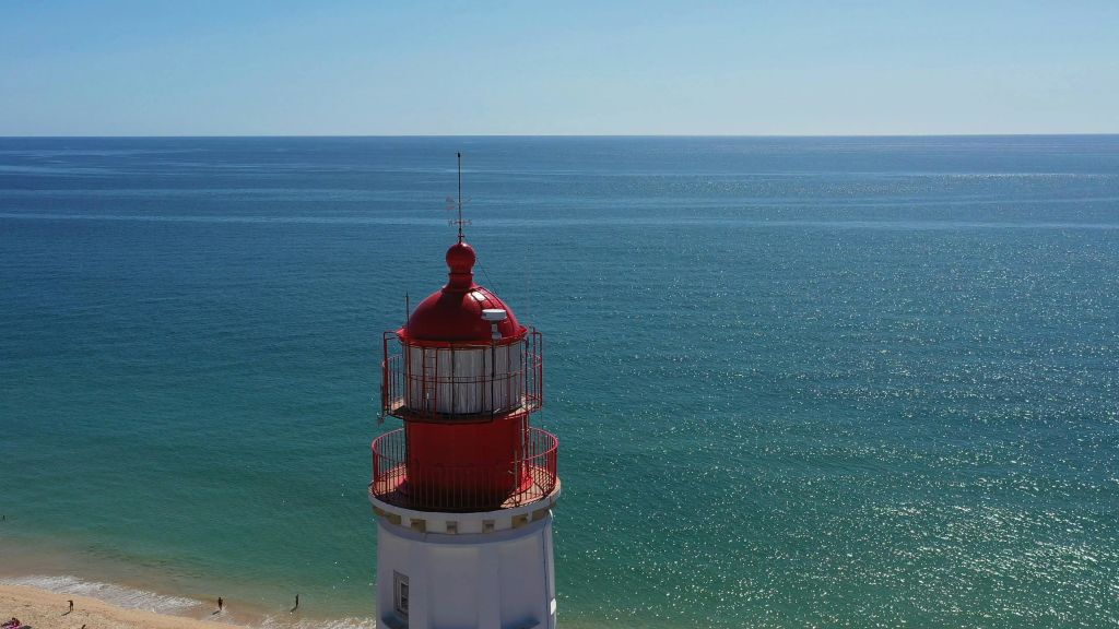 맑고 푸른 하늘 아래 고요하고 넓은 바다가 내려다보이는 빨간색과 흰색 등대. 기슭에 모래 해변이 보입니다.