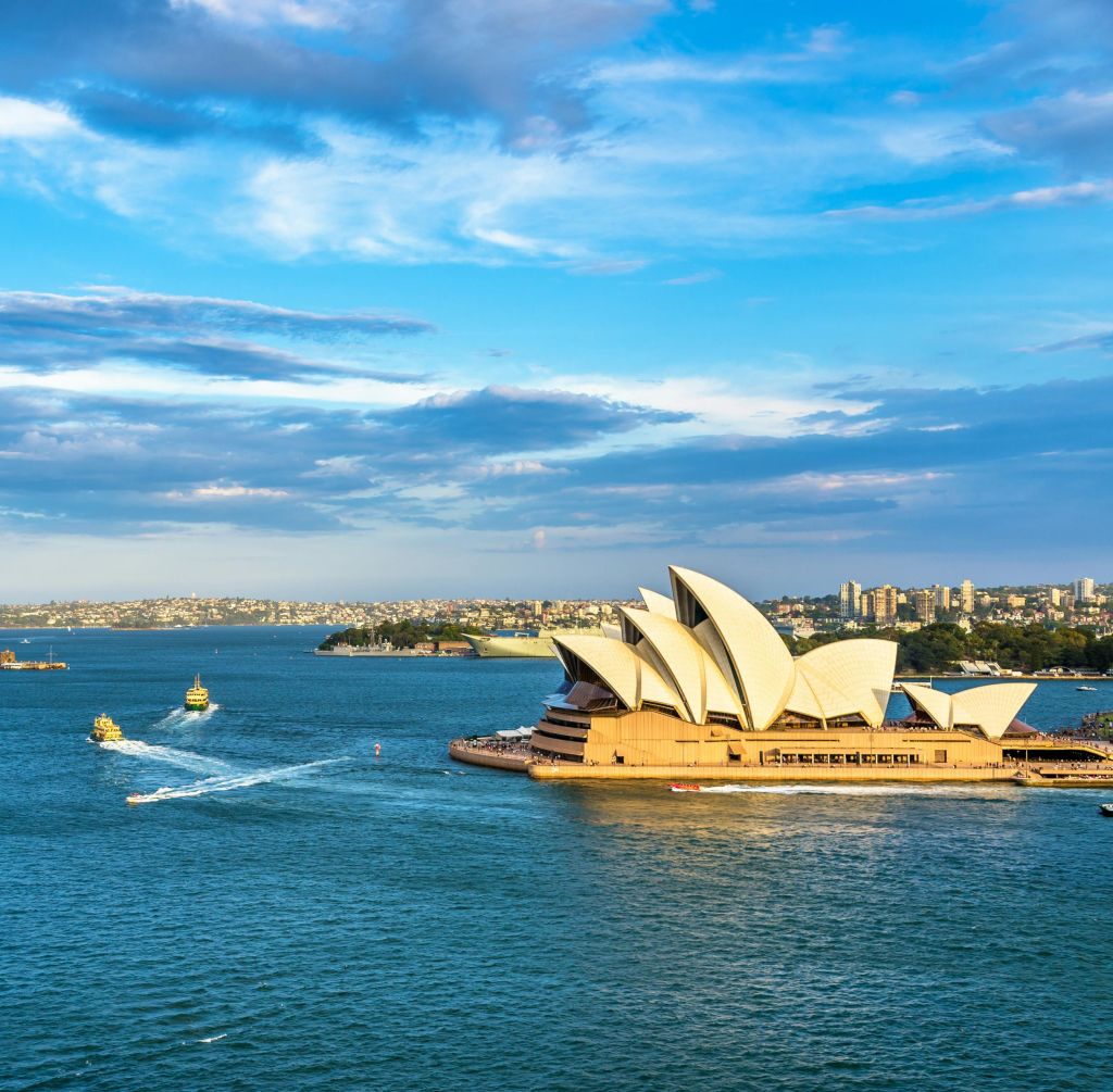 시드니 오페라 하우스는 독특한 돛을 달고, 보트가 있는 물로 둘러싸여 있으며, 부분적으로 흐린 하늘 아래에서 도시 풍경을 감상할 수 있습니다.