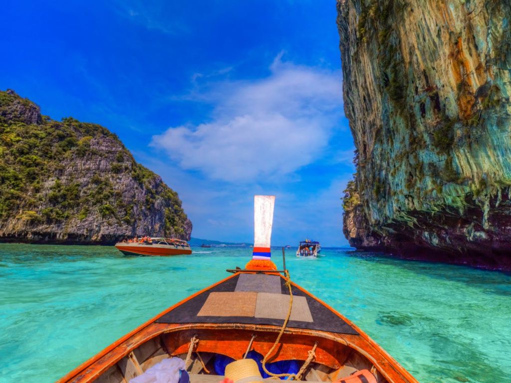 높은 절벽으로 둘러싸인 맑은 청록색 바다의 나무 보트와 밝고 푸른 하늘 아래 다른 보트에서 바라보는 풍경.