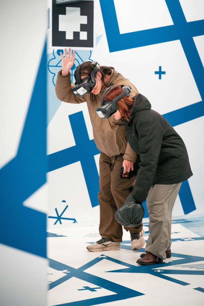 파란색과 흰색의 기하학적 패턴이 있는 방에서 가상 현실 헤드셋을 착용한 두 사람이 상호 작용합니다.