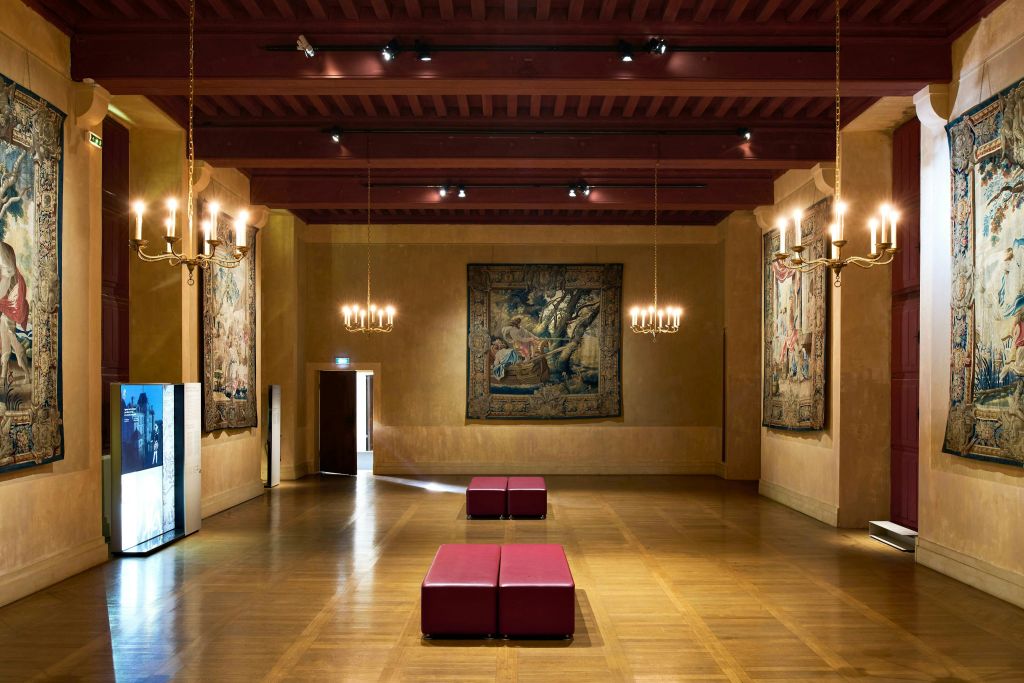 벽에 나무 바닥, 빨간 벤치, 걸이식 샹들리에, 장식용 태피스트리가 있는 넓은 객실입니다.