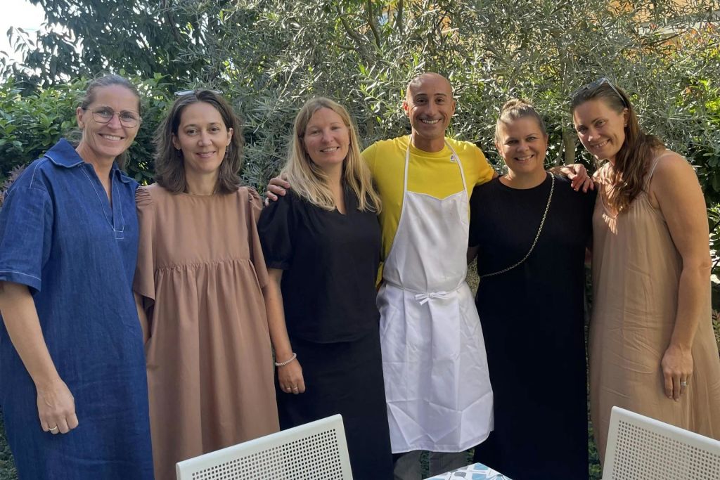 5인 그룹이 야외에 서서 미소를 짓고 있습니다. 노란 셔츠를 입은 한 사람이 앞치마를 착용하고 있습니다. 나무와 야외 의자가 보입니다.