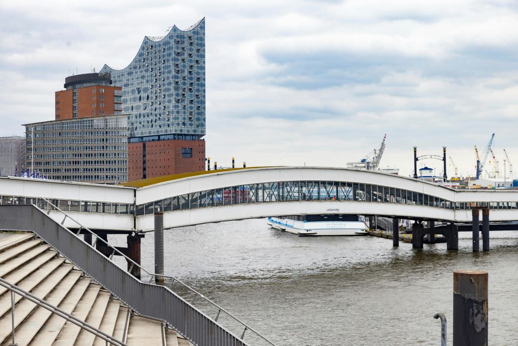 물결 모양의 지붕, 강 위의 보행자 다리, 계단, 흐린 하늘 아래 정박된 보트가 있는 현대적인 건물입니다.