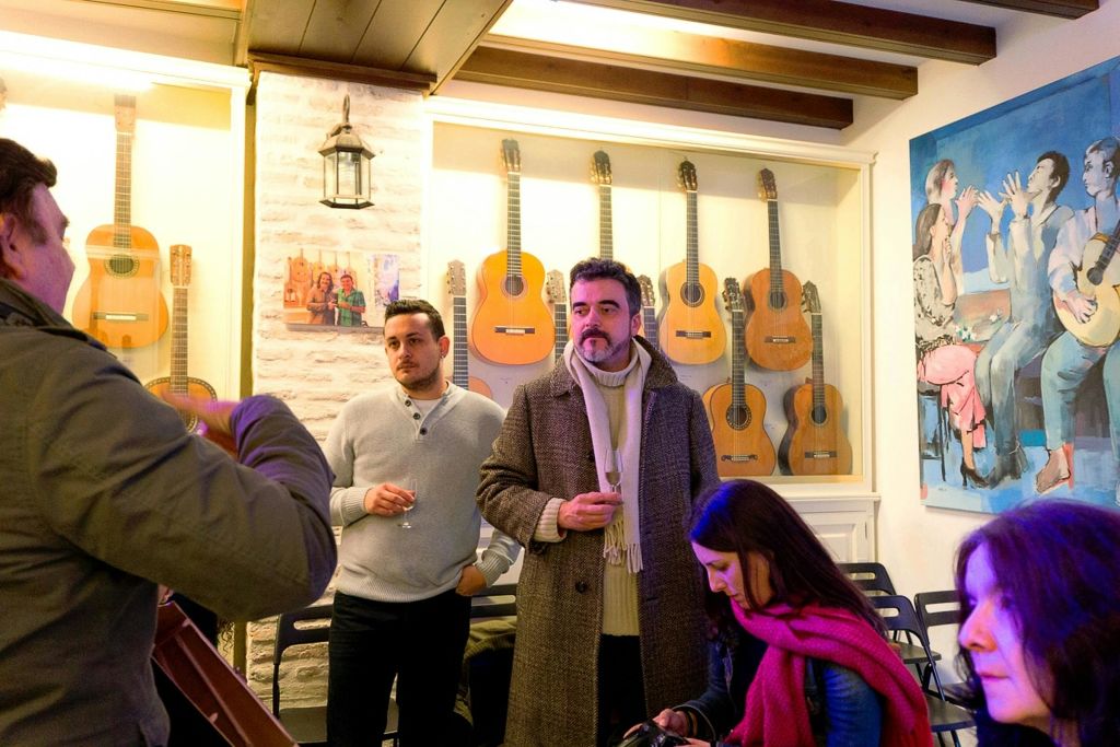 벽에 여러 대의 기타가 걸려 있고 커다란 그림을 배경으로 방에 서서 앉아 있는 사람들.