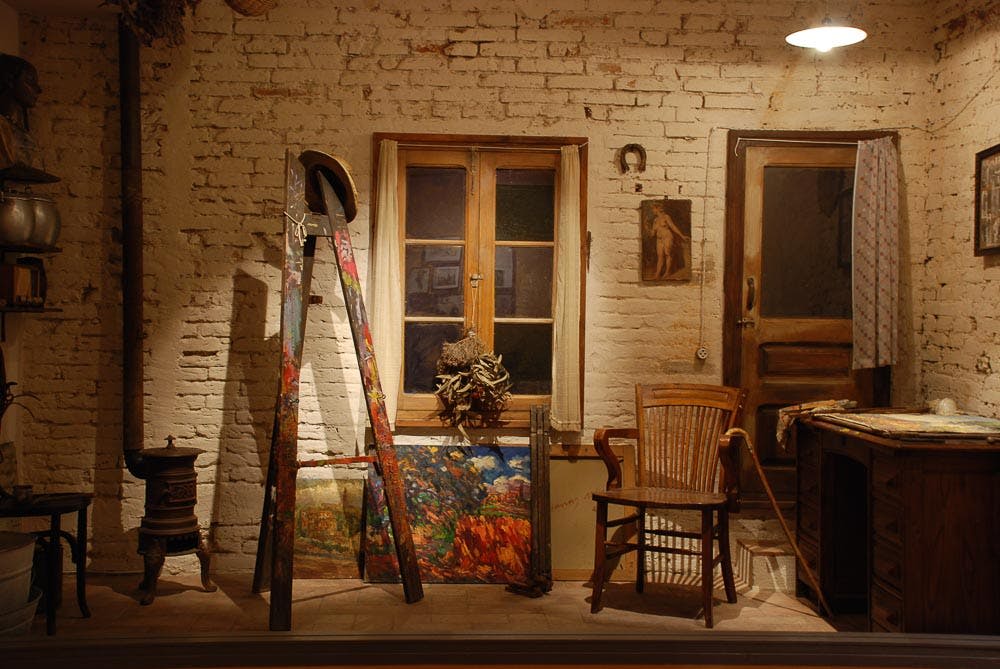 페인트가 흩뿌려진 이젤, 액자에 담긴 그림, 나무 의자, 벽돌 벽에 걸린 창문이 있는 작가의 작업실입니다.