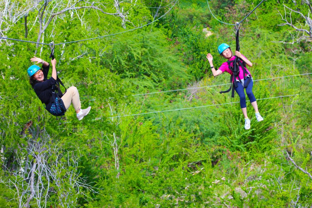 울창한 푸른 숲을 가로지르는 두 사람이 헬멧과 안전 장비를 착용한 채 손을 흔들고 있습니다.
