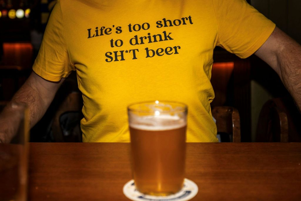 "SH \* T 맥주를 마시기에는 인생이 너무 짧다" 라는 문구가 적힌 노란색 셔츠와 앞쪽 테이블에 놓인 맥주 한 잔.