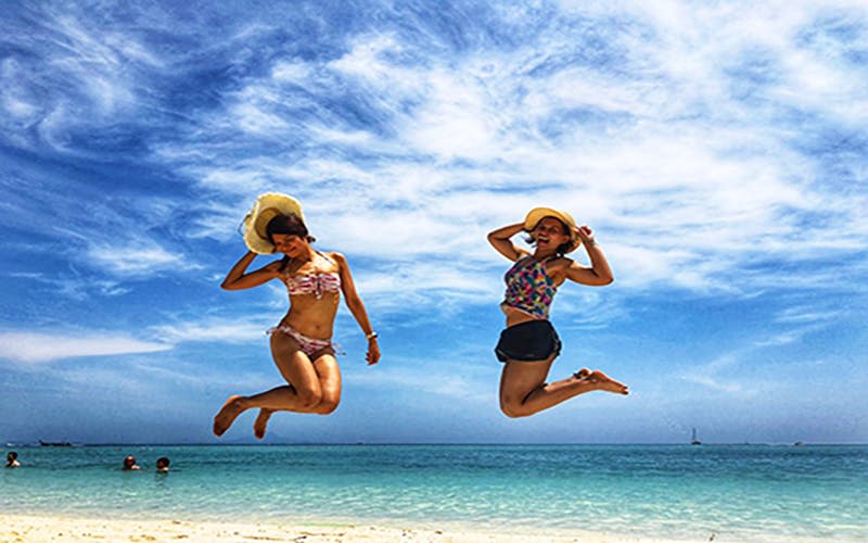 맑고 푸른 물과 부분적으로 흐린 하늘을 배경으로 수영복과 모자를 입은 두 여성이 공중에서 뛰어내리고 있습니다.