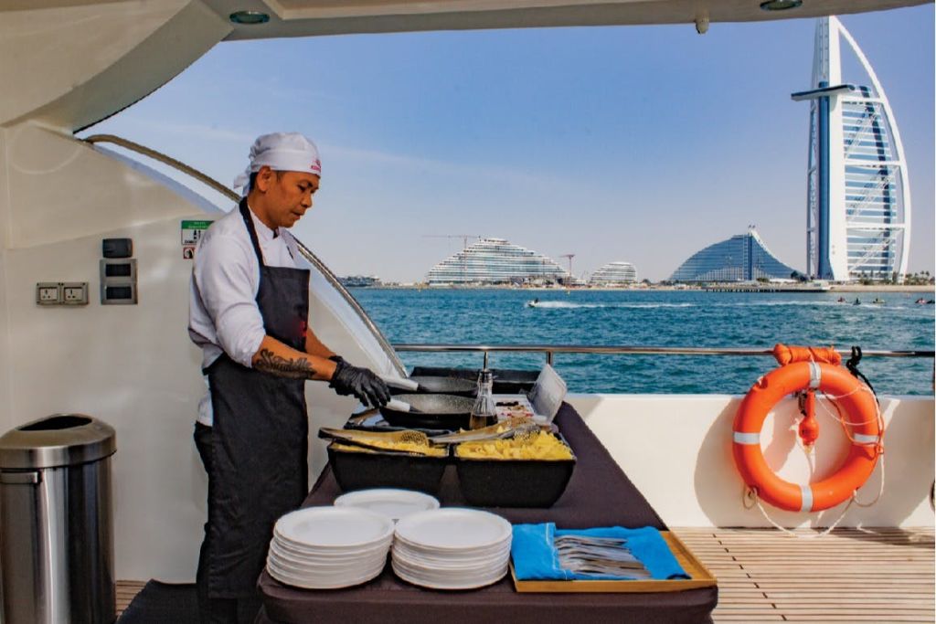 흰색 모자와 앞치마를 두른 셰프가 바다와 현대적인 건물을 배경으로 보트를 타고 음식을 준비합니다.