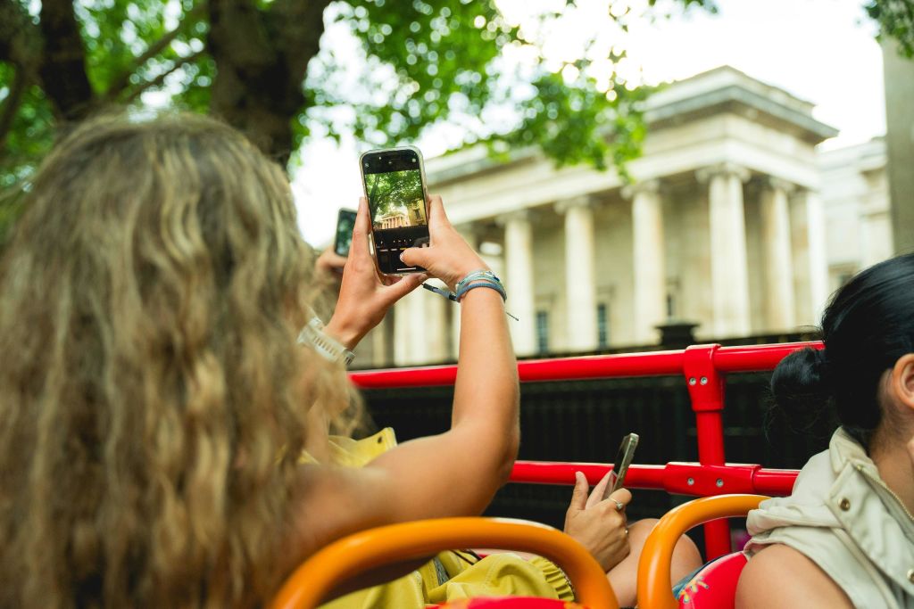 빨간색 오픈탑 차량에 앉아 스마트폰으로 기둥이 있는 건물의 사진을 찍는 사람.