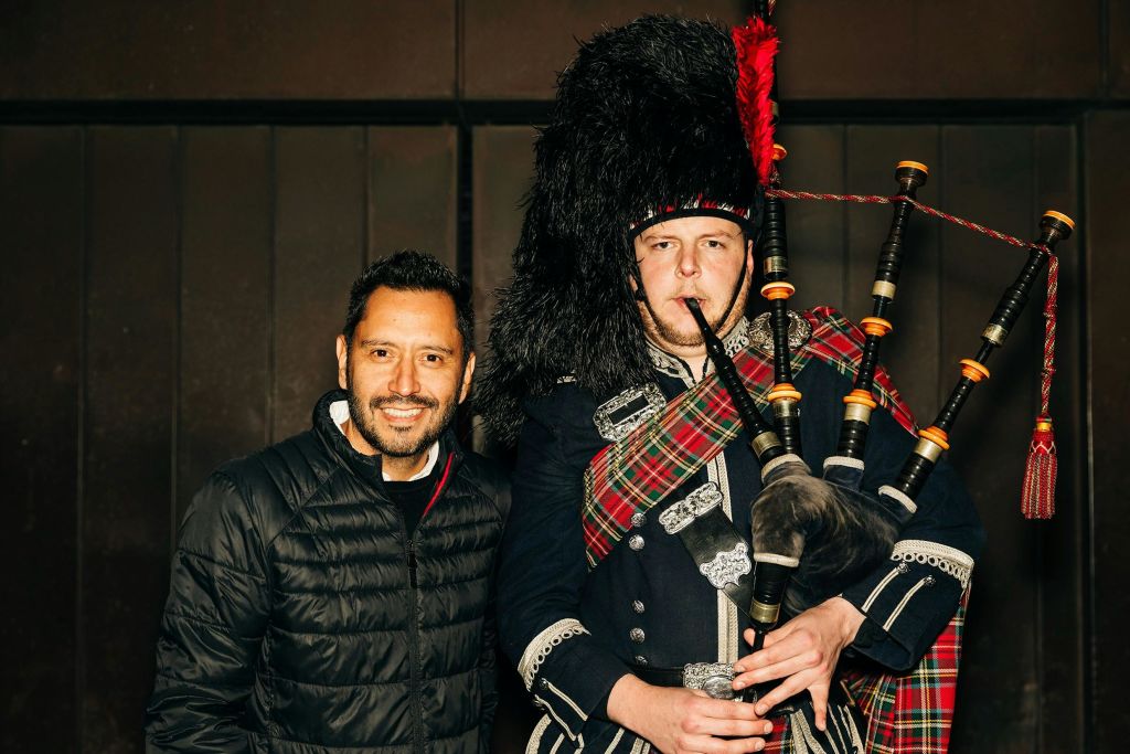 실내에서 깃털 달린 모자, 격자 무늬 띠 등 스코틀랜드 전통 복장을 한 백파이퍼 옆에서 웃고 있는 남성.