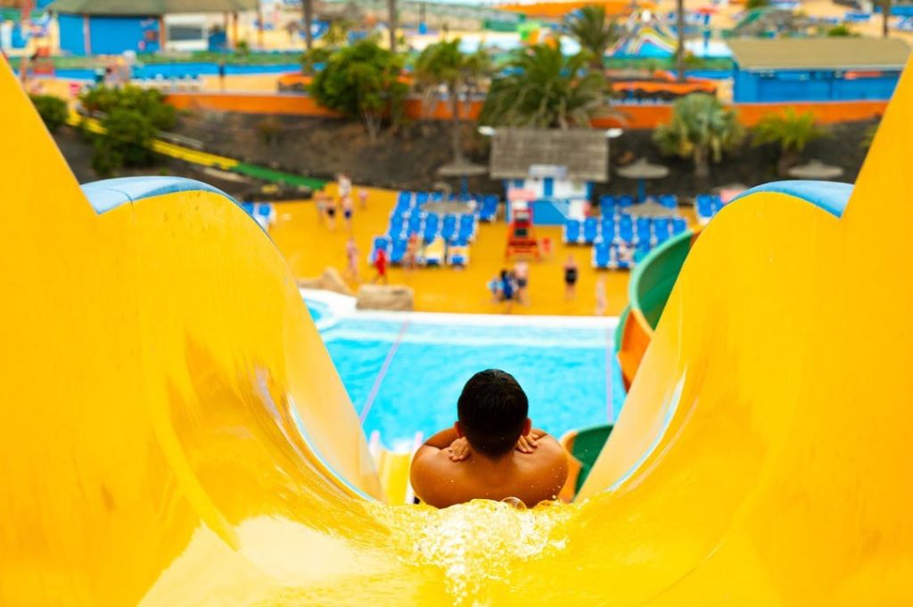 수영장과 의자, 야자수를 배경으로 워터파크에서 노란색 미끄럼틀을 타고 내려오는 사람.