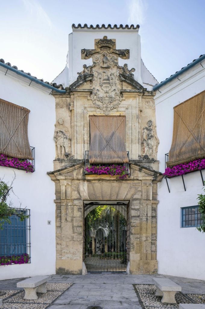 화려한 조각, 닫힌 나무 셔터, 빗장 문, 활기찬 보라색 꽃이 있는 건물의 유서 깊은 석조 입구.