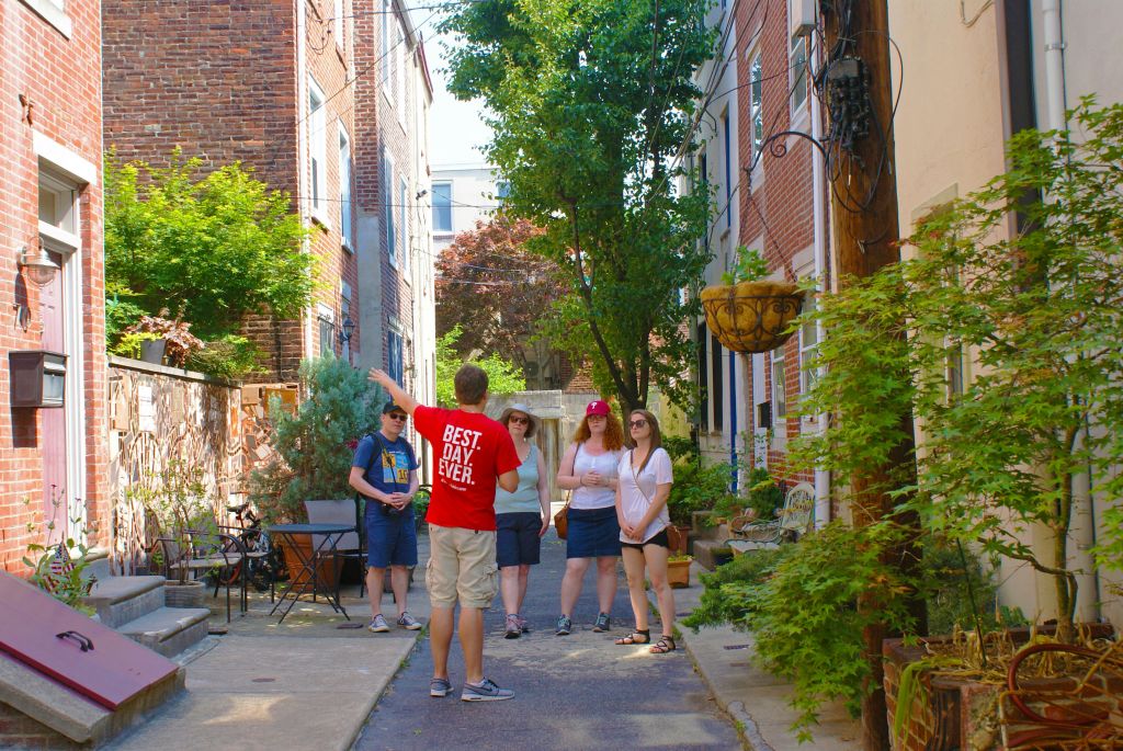 한 무리의 사람들이 빨간 셔츠를 입은 투어 가이드가 무언가를 가리키며 골목길에 서 있습니다. 벽돌 건물과 녹지가 주변을 둘러싸고 있습니다.