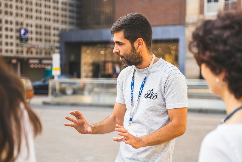 회색 셔츠를 입고 랜야드를 든 남성이 야외에서 제스처를 취하고 있습니다.