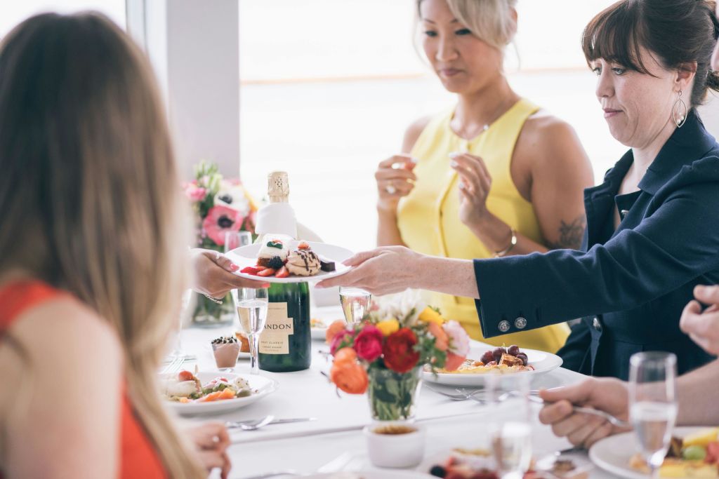 식탁에 앉은 두 명 중 한 명은 노란색 드레스를 입고 한 명은 어두운색 드레스를 입고 있습니다. 식탁에는 음식, 샴페인, 꽃이 준비되어 있습니다.