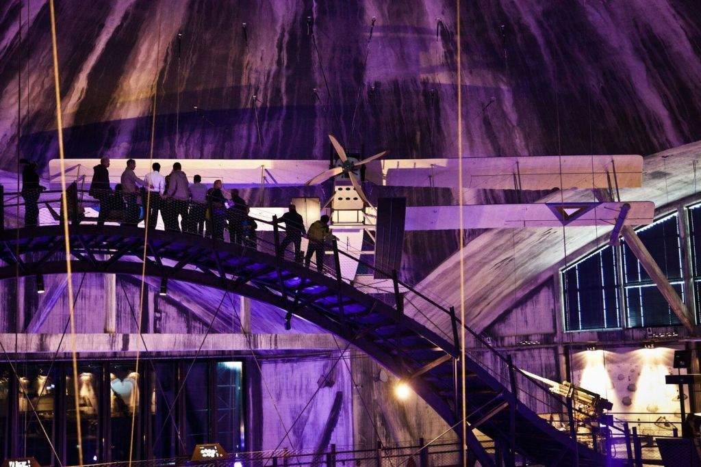 사람들은 보라색 조명이 있는 조명이 희미한 산업 구조물 내부의 높고 곡선된 다리를 걷습니다.