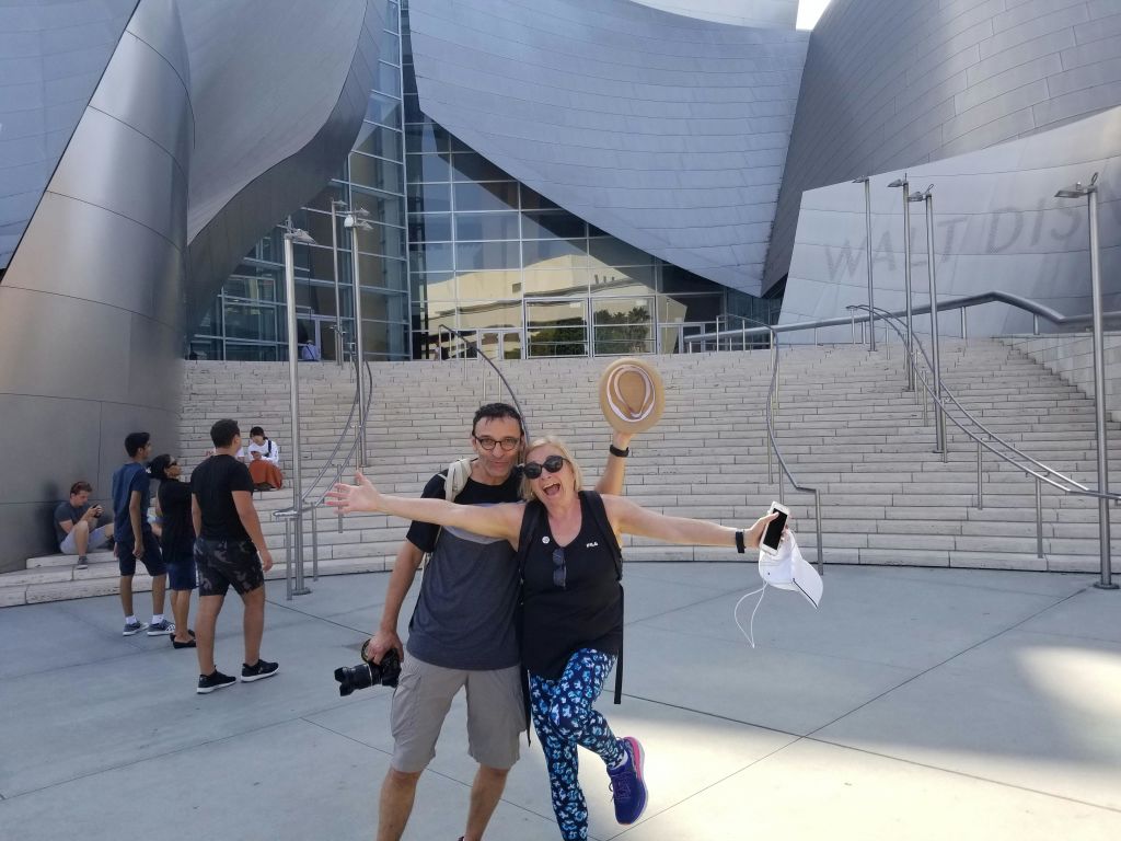 캐주얼한 복장을 한 두 사람이 곡선형 금속 건축물이 있는 현대적인 건물의 계단에서 즐거운 포즈를 취하고 있습니다. 배경에 있는 다른 사람들.