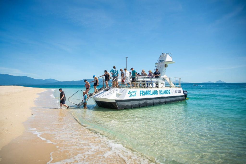 한 무리의 사람들이 "프랭클랜드 제도" 라는 이름의 보트를 타고 맑고 푸른 물이 흐르는 햇살 아래 모래 해변으로 내립니다.