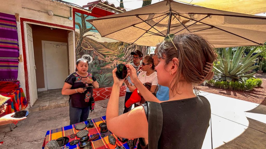 한 여성이 우산 아래 다채로운 벽화와 물품 테이블이 있는 건물 옆에서 작은 그룹에게 물건을 보여주고 있습니다.
