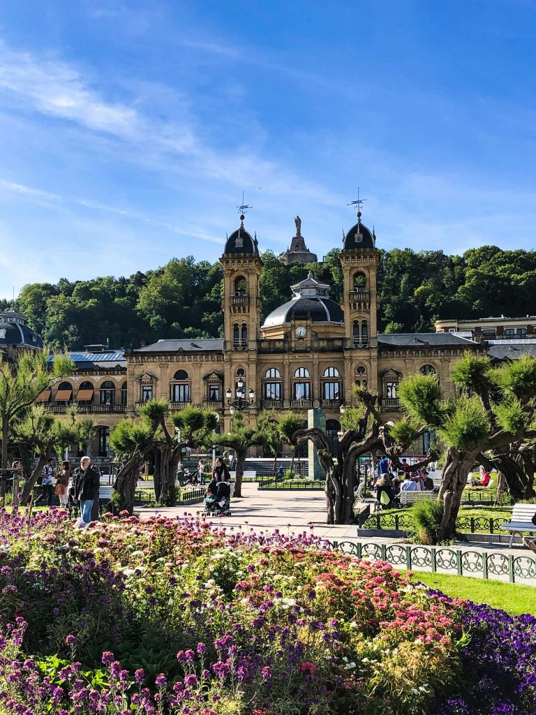 언덕을 배경으로 맑고 푸른 하늘 아래 트윈 타워, 정원, 벤치에 앉아 걷거나 앉아 있는 사람들이 있는 유서 깊은 건물입니다.
