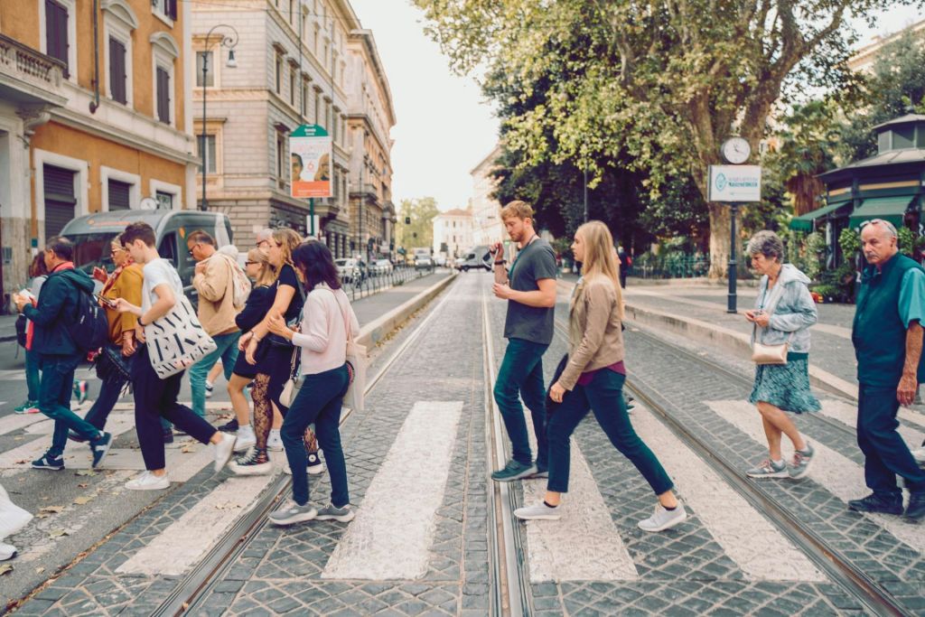 건물, 자전거, 시계 디스플레이로 둘러싸인 얼룩말 교차로에서 가로수가 늘어선 거리를 가로지르는 사람들.