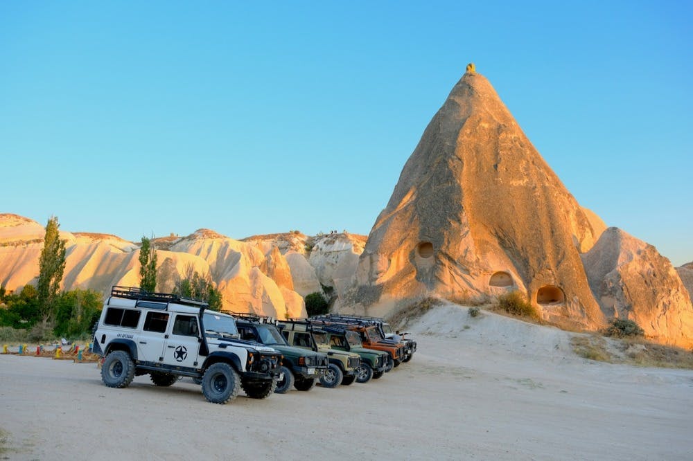 맑고 푸른 하늘 아래 동굴 같은 개구부가 있는 바위 지층 옆에 주차된 오프로드 차량이 줄지어 있습니다.