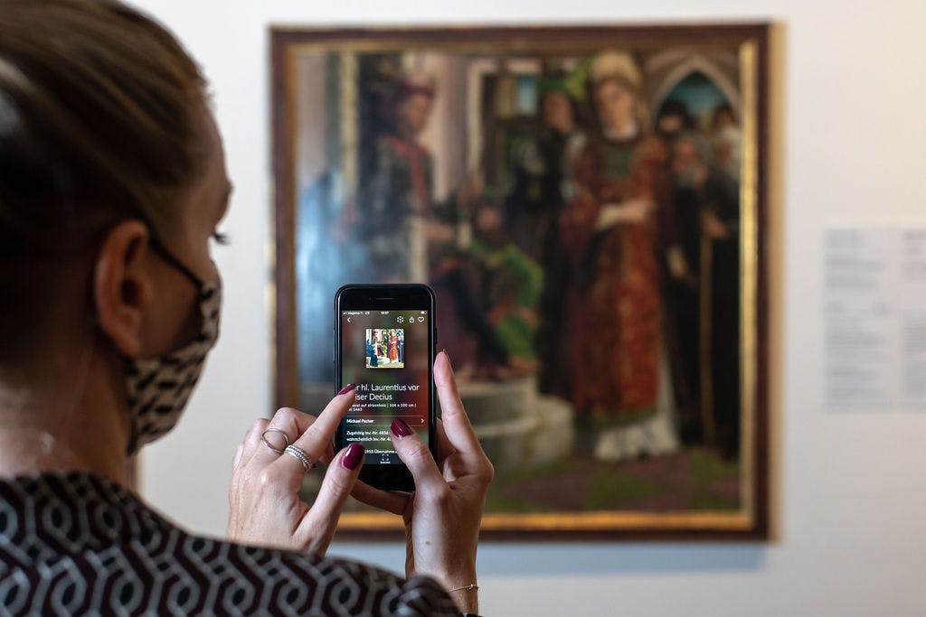 스마트폰을 들고 있는 사람이 박물관에서 액자에 담긴 작품의 사진을 찍고 있습니다. 작품에 대한 텍스트 세부 정보는 휴대전화 화면에서 확인할 수 있습니다.