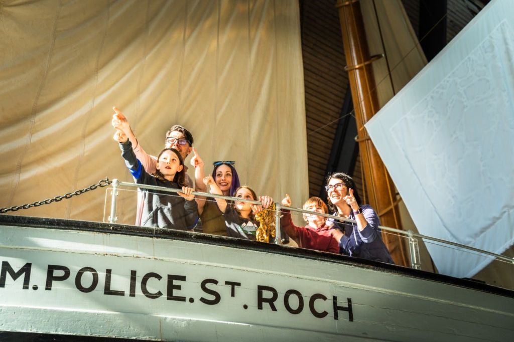 한 무리의 사람들이 "Police St. Roch" 라는 이름의 배 갑판에 서서 밝은 실내 환경에서 미소를 짓고 있습니다.