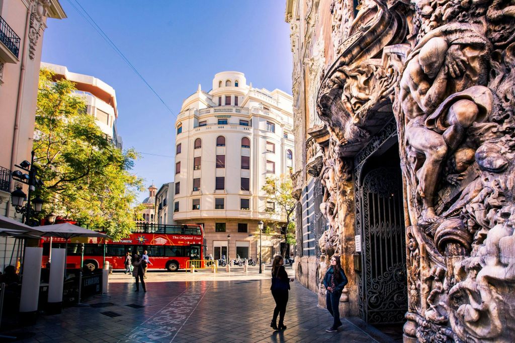 화려한 건물 외관, 빨간색 2층 투어 버스, 산책하는 사람들, 둥근 모퉁이 건물을 배경으로 한 도심의 거리.