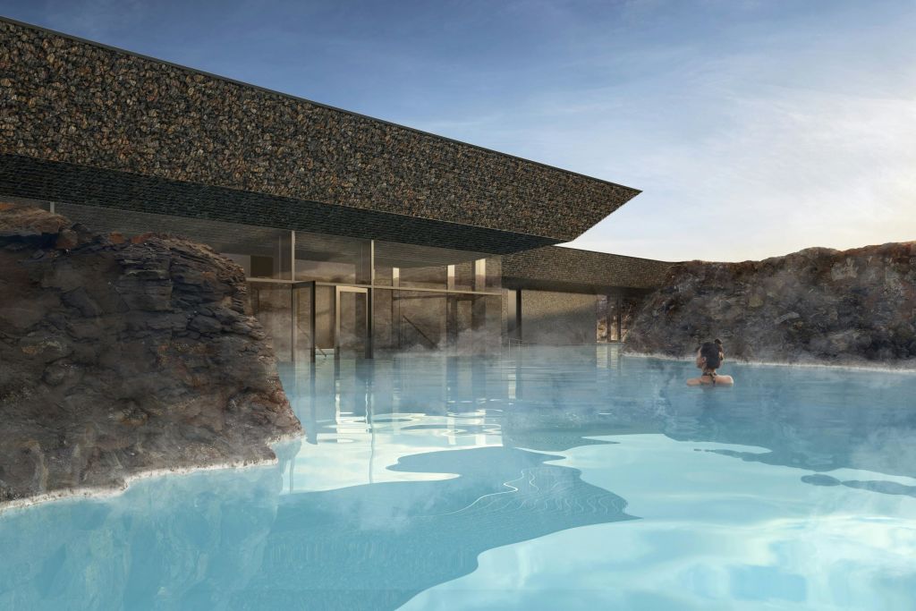 바위 벽이 있는 현대적인 건물로 고요한 푸른 지열 수영장이 내려다보입니다. 한 사람이 맑은 하늘 아래에서 물속을 헤엄치고 있습니다.