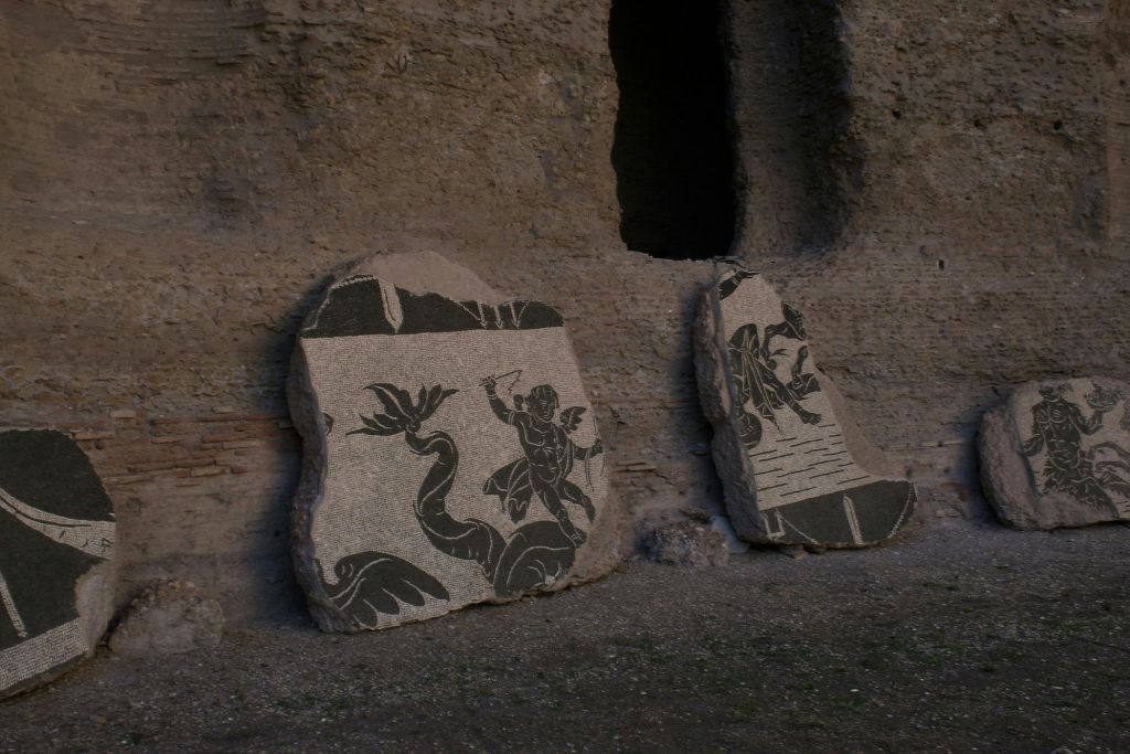 거친 돌담에 기댄 흑백 디자인의 고대 석조 모자이크 두 개. 하나는 뱀을 가진 사람을 묘사합니다.