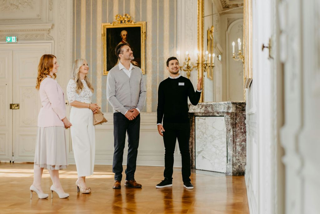 초상화와 샹들리에가 있는 우아한 객실에서 성인 3명과 대화하는 동안 투어 가이드가 제스처를 취합니다.