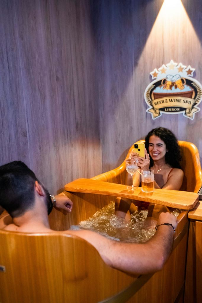스파에서 나무로 만든 온수 욕조에서 음료를 마시며 휴식을 취하는 두 사람. 벽에 'Beer & Wine Spa Lisbon' 이라는 문구가 적혀 있습니다.