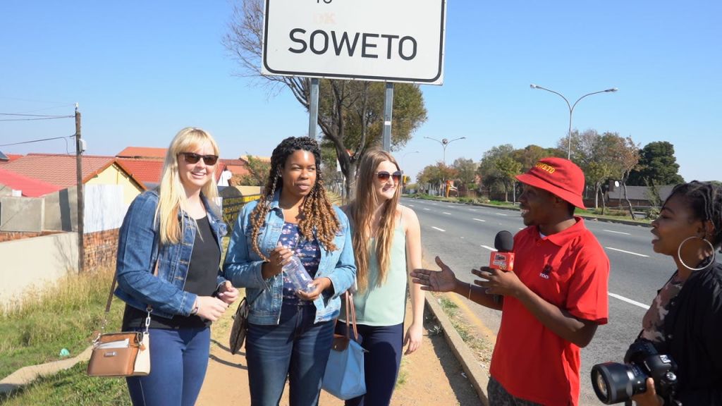 주택과 나무를 배경으로 '소웨토' 라고 적힌 거리 표지판을 배경으로 세 명의 여성이 서서 이야기를 나누고 있습니다.