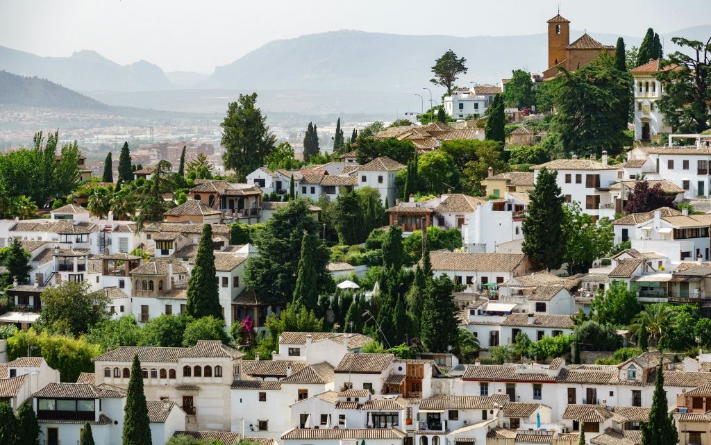 흰색 건물, 타일 지붕, 울창한 녹지가 있는 언덕 마을입니다. 맑은 하늘 아래 먼 산맥이 보인다.