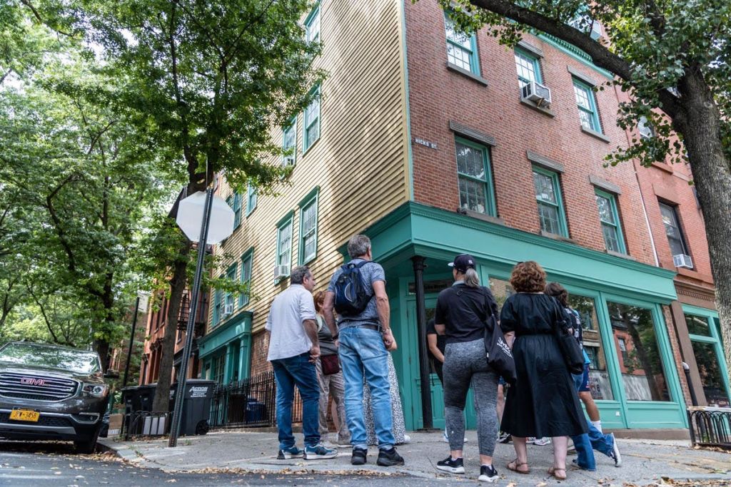 한 무리의 사람들이 길 모퉁이에 서서 벽돌과 나무 사이딩이 있는 2층 건물을 바라보고 있습니다.