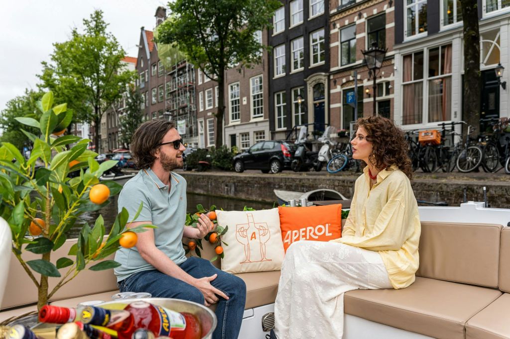 두 사람이 보트에 앉아 이야기를 나누고 있습니다. 그 뒤편에는 주차된 자동차와 자전거가 있는 운하와 연립주택이 있습니다.