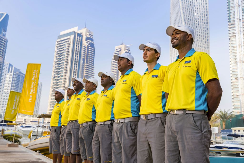 맑은 날, 노란 셔츠와 회색 반바지를 입은 여덟 명의 유니폼을 입은 일꾼들이 높은 건물 앞에 서 있습니다.