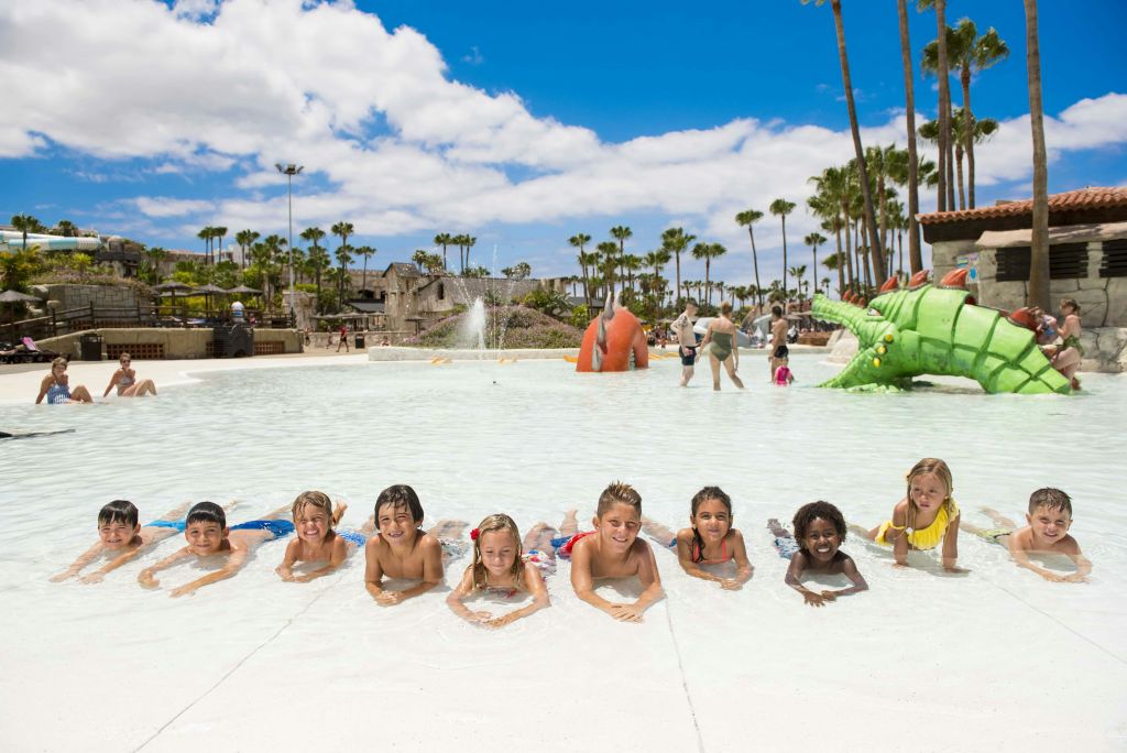 장난기 가득한 물놀이 공간과 야자수를 배경으로 얕은 수영장에서 배를 깔고 누워 있는 어린이들.