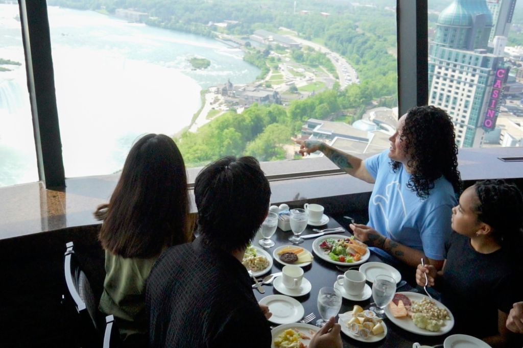 강, 녹지, 건물의 경치가 내려다보이는 커다란 창문이 있는 레스토랑에서 세 사람이 식사를 하고 있습니다.