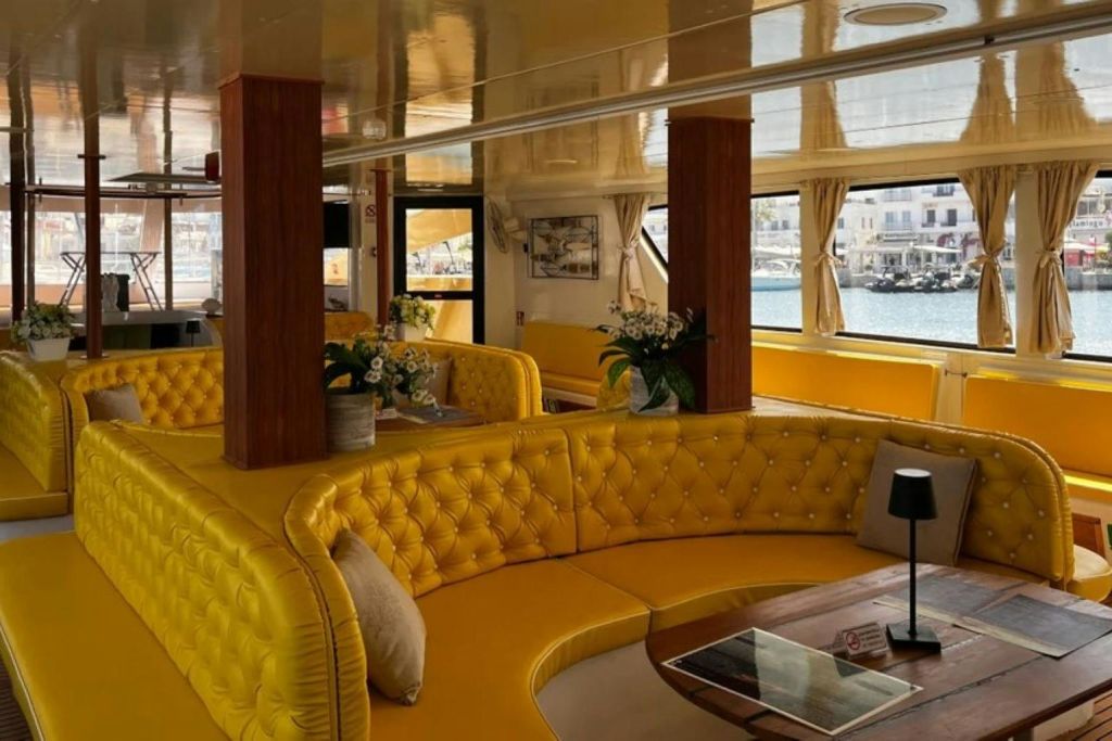 노란색 터프팅 소파, 목재 액센트, 화분, 물과 정박지가 내려다보이는 커다란 창문이 있는 아늑한 좌석 공간.