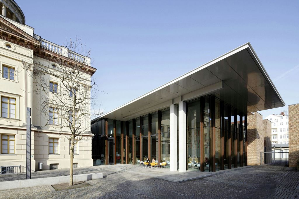 화창한 날, 오래된 석조 건물에 인접한 넓은 돌무더기 아래 야외 좌석이 있는 현대적인 유리 전면 건물.