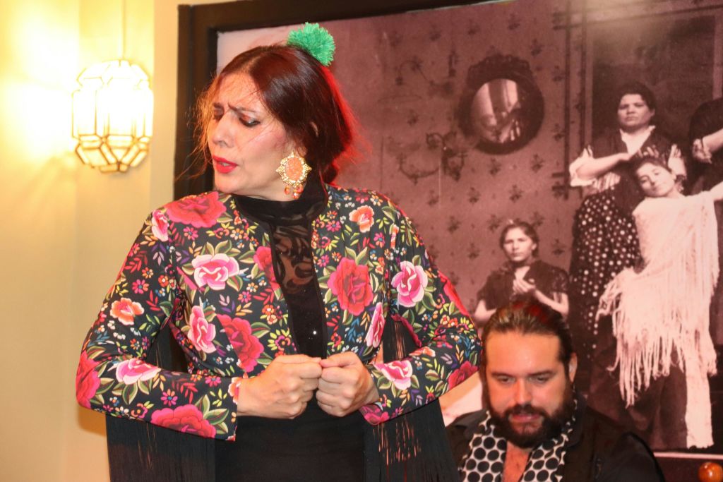 꽃무늬 숄을 입은 여성이 앉아 있는 남성 옆에서 빈티지한 흑백 사진을 배경으로 공연을 펼치고 있습니다.