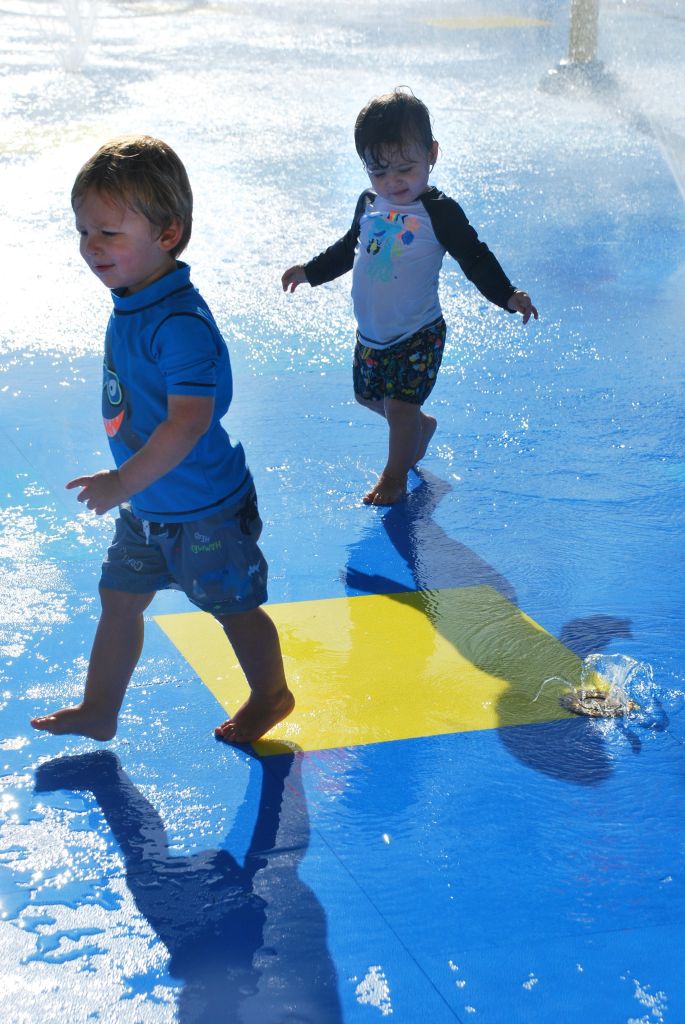 두 어린이가 워터파크에서 물 스프레이로 인한 안개로 둘러싸인 파란색과 노란색 젖은 표면을 걷고 있습니다.