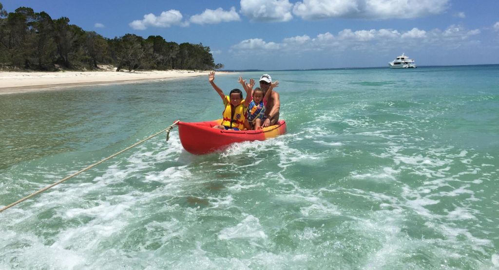 모래 해변 근처의 맑고 얕은 물 위에서 세 사람이 작은 빨간 보트를 타고 구명조끼를 흔들고 입고 있습니다.