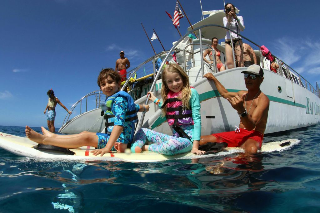 다른 사람들과 함께 보트 근처의 물속에서 서핑 보드에 앉아있는 구명 조끼를 입은 두 명의 어린이.